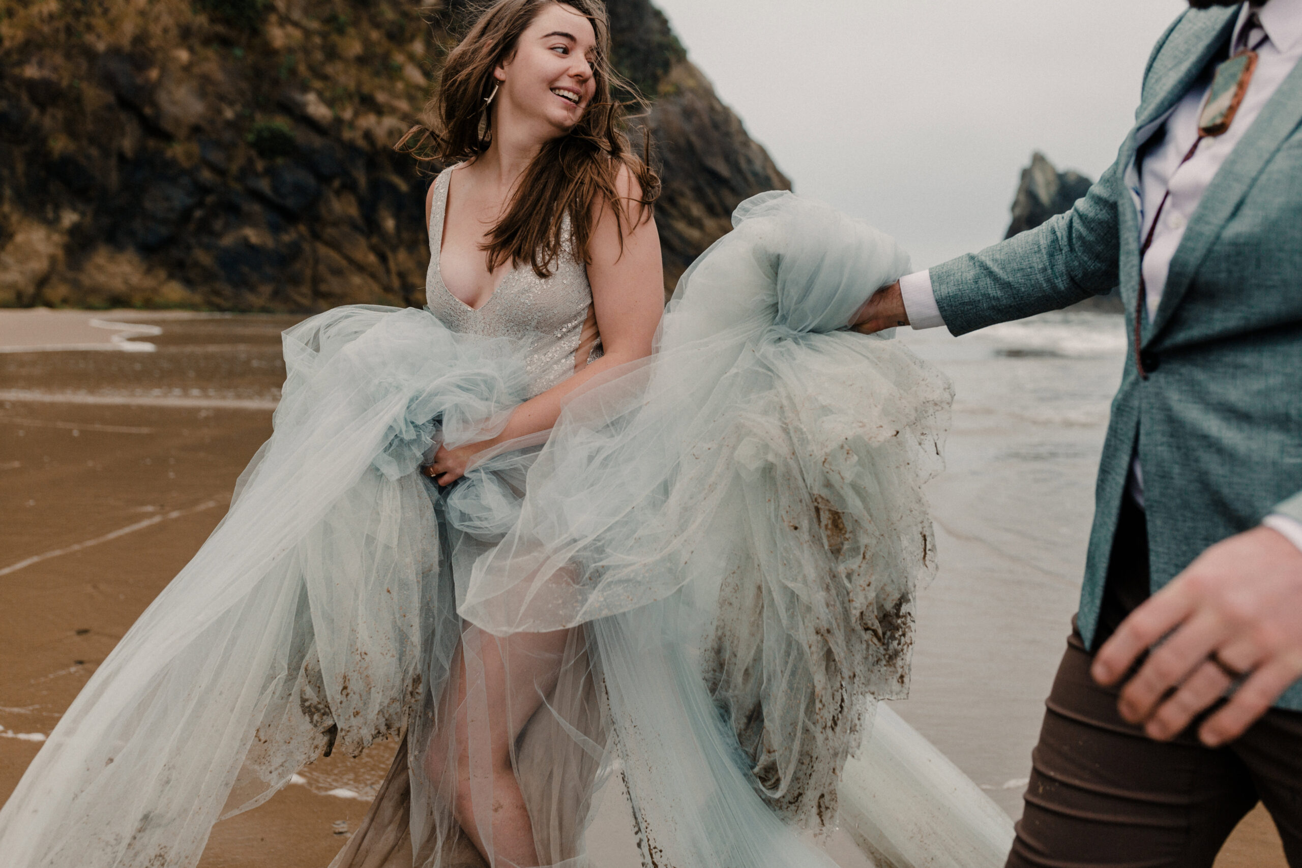 bride running from waves as groom helps hold dress during beach elopement